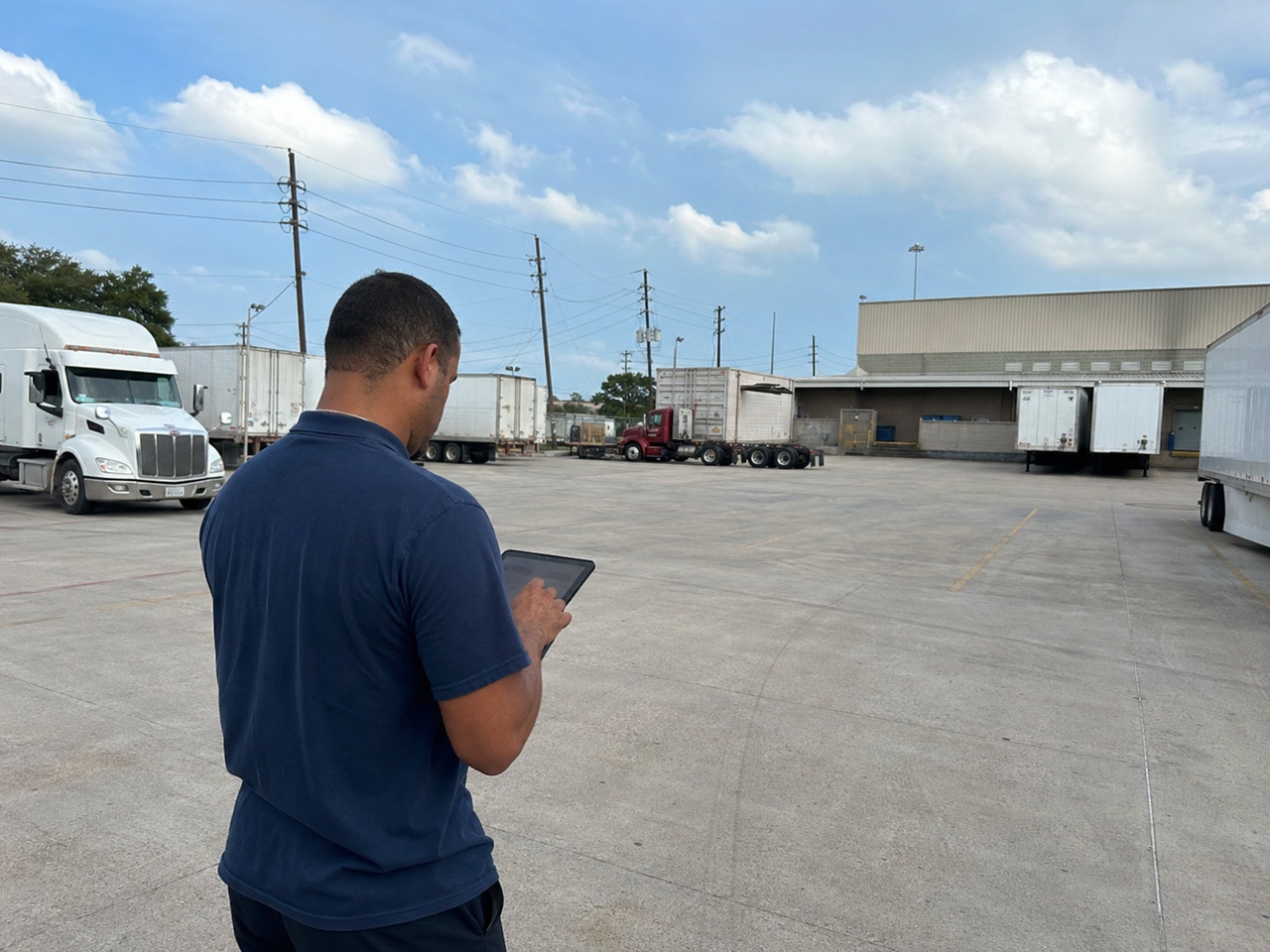 A freight broker stands in a Texas commercial trucking yard holding a tablet while multiple semi-trucks and trailers are staged near a warehouse loading dock behind him, depicting the role a freight broker plays in coordinating carrier assignments and the potential liability that can arise in a Texas truck accident when a broker selects an unqualified or unsafe motor carrier.